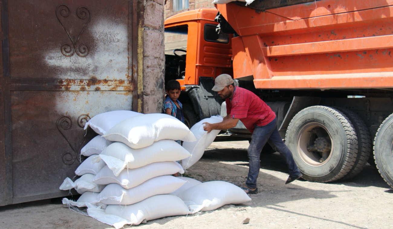 A man in a red shirt unloads white bags from a large orange truck while a small child looks on. 