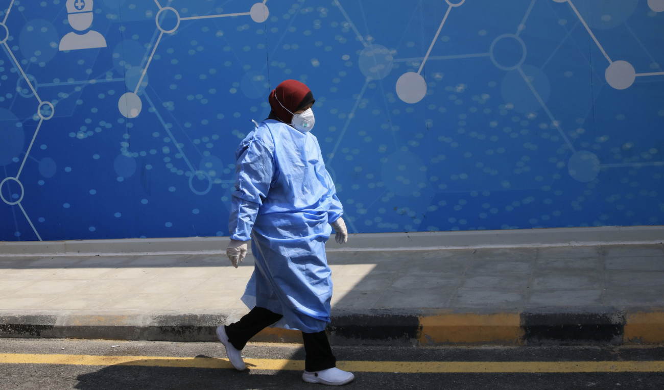 Une infirmière vêtue d'une blouse médicale bleue et portant un foulard autour de la tête ainsi qu'un masque de protection respiratoire et des gants de protection marche dans une allée le long d'un bâtiment à la façade bleue.