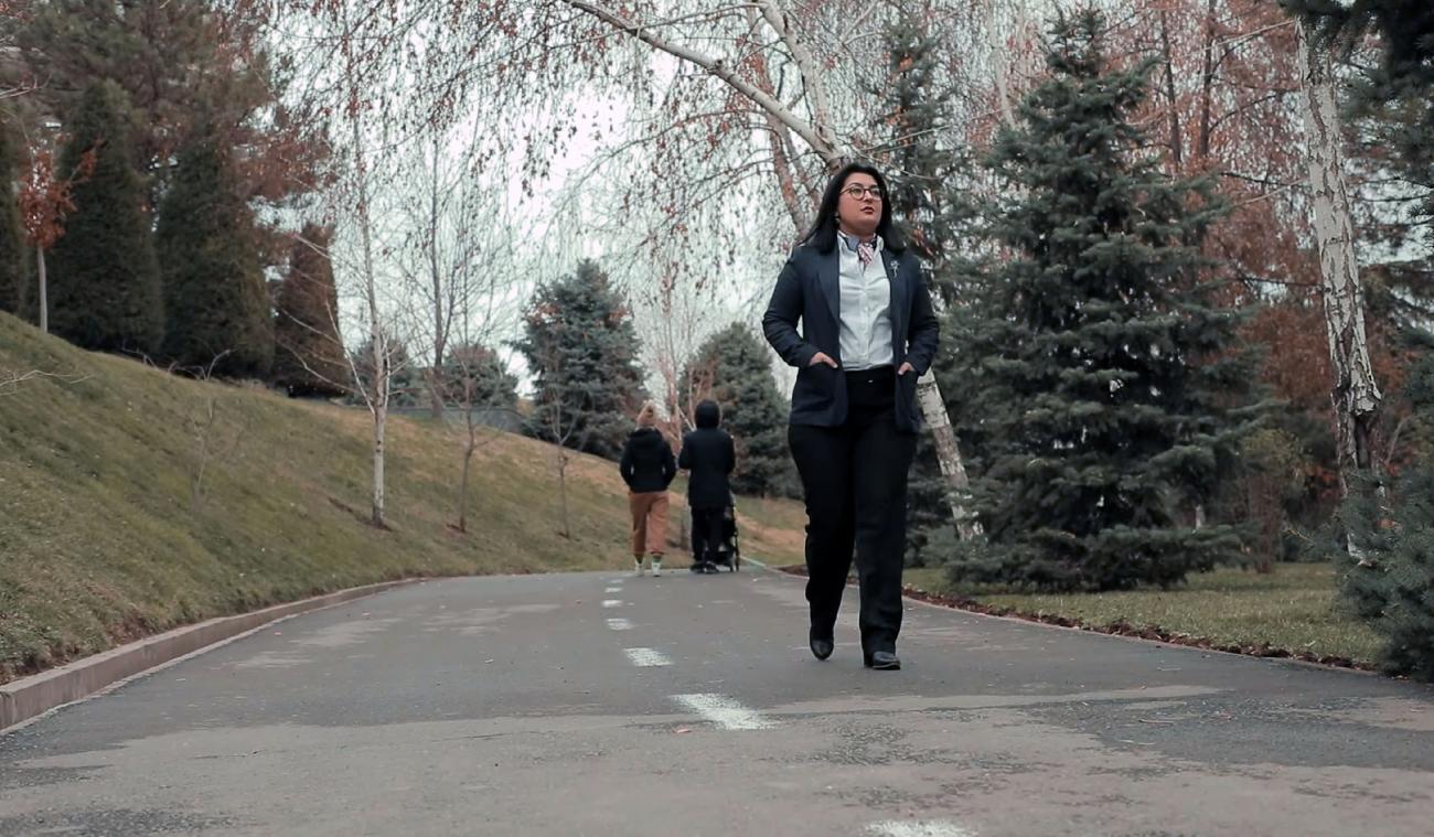 Aqida Mokhirova camina pensativa por la vía asfaltada de un parque, mientras en el fondo dos personas, que vemos de espaldas y una de las cuales empuja un cochecito de bebé, se alejan de ella caminando en la dirección contraria.