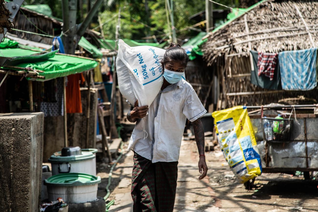 A person in a face mask carries a large white sack on their shoulder down a street with many homes. 