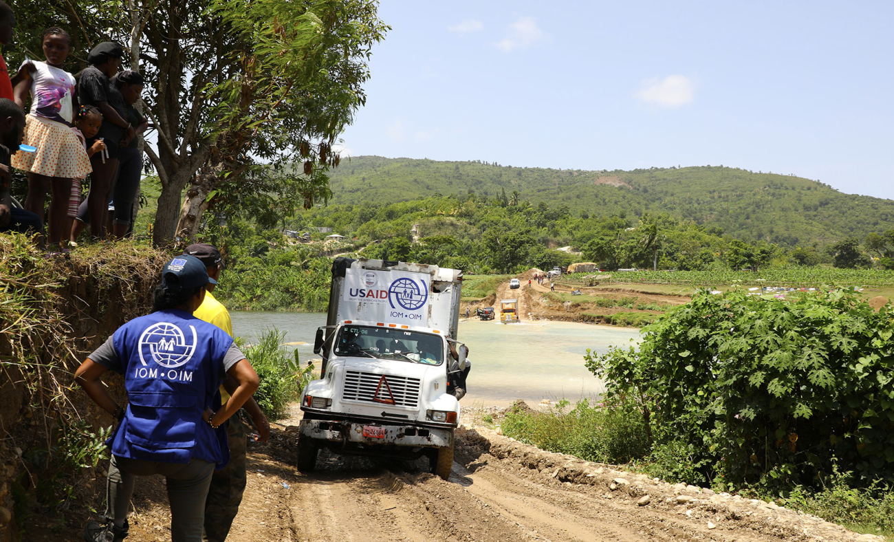 IOM workers stand on the side of the road as a large truck drives down the dirt road and children look down from above.