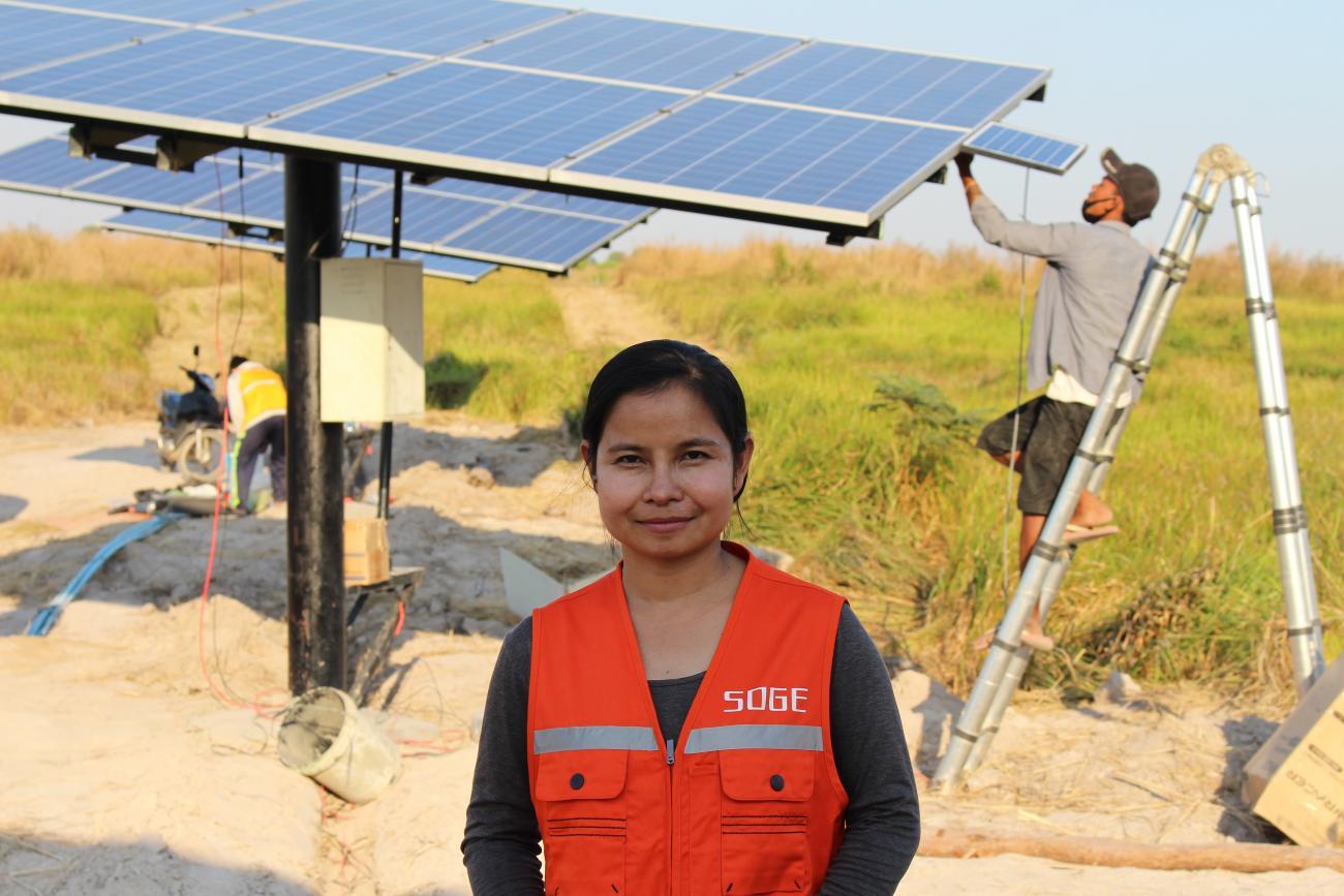 A woman in an orange vest smiles in front of large solar panels. 