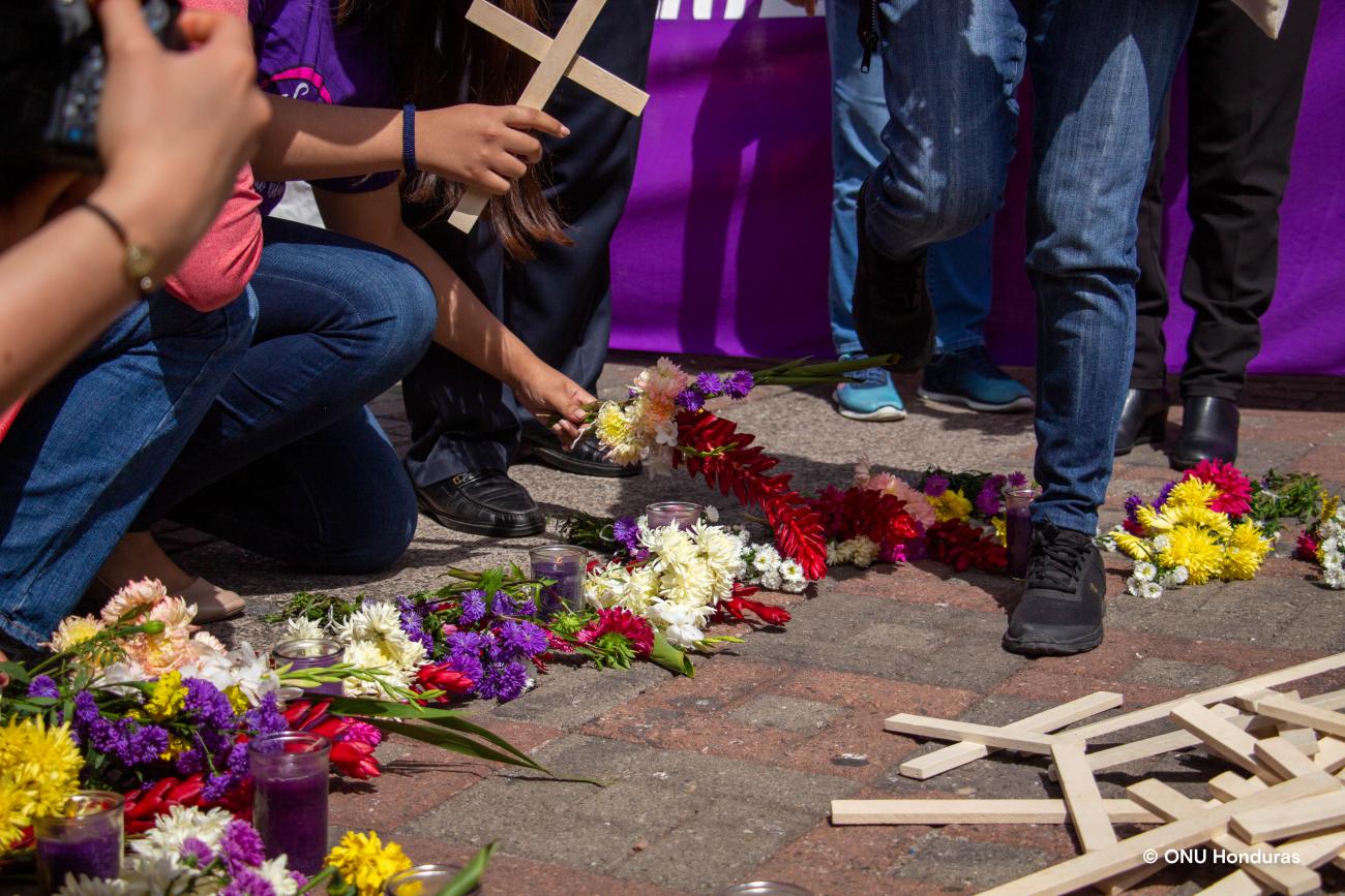 La imagen muestra los brazos de las personas que sostienen cruces arrodilladas junto a flores y más cruces amontonadas.