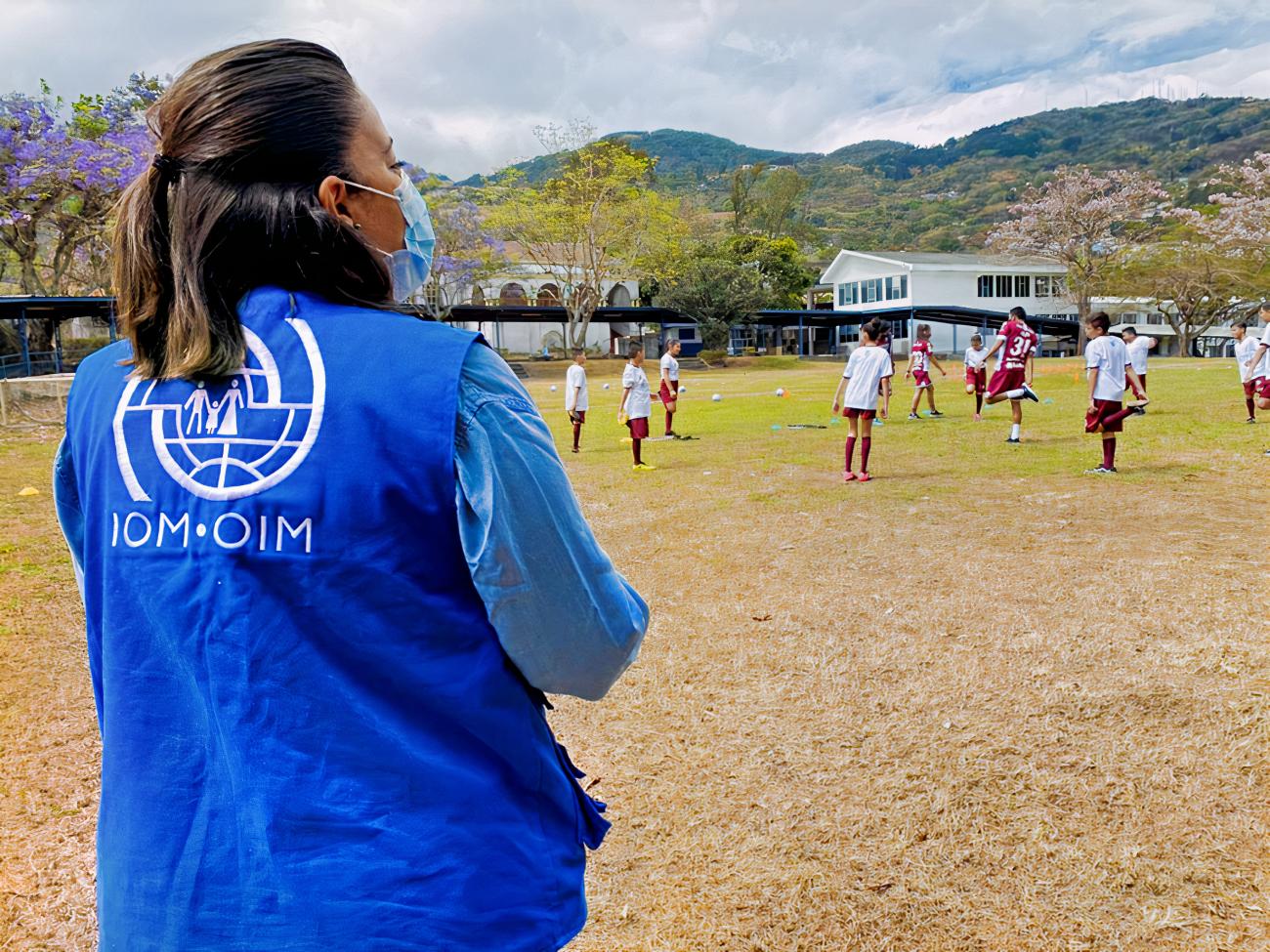 A woman in an IOM vest looks onto a soccer pitch with many kids playing soccer. 