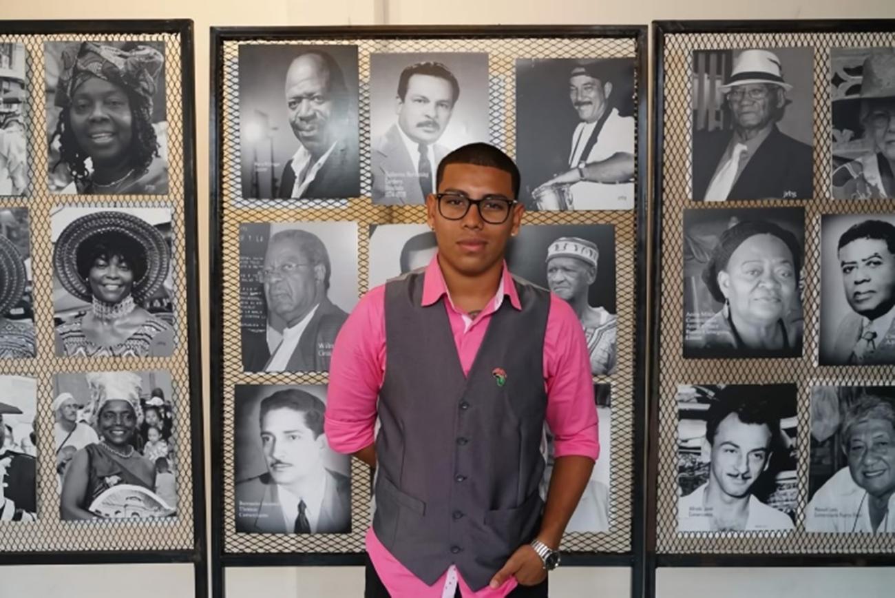 Un joven se encuentra frente a una pared llena de retratos en blanco y negro.