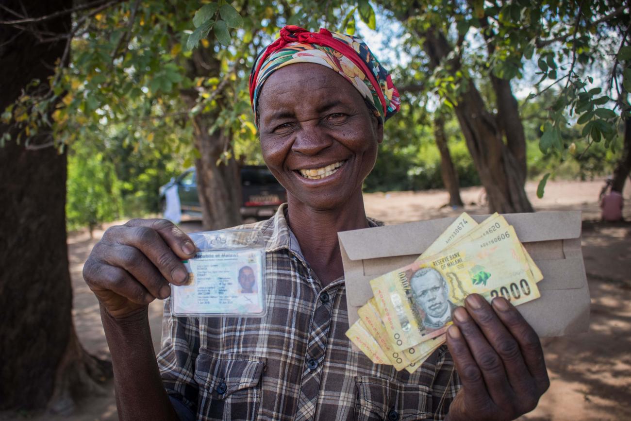 A smiling woman holds up cash assistance and her identification card. 