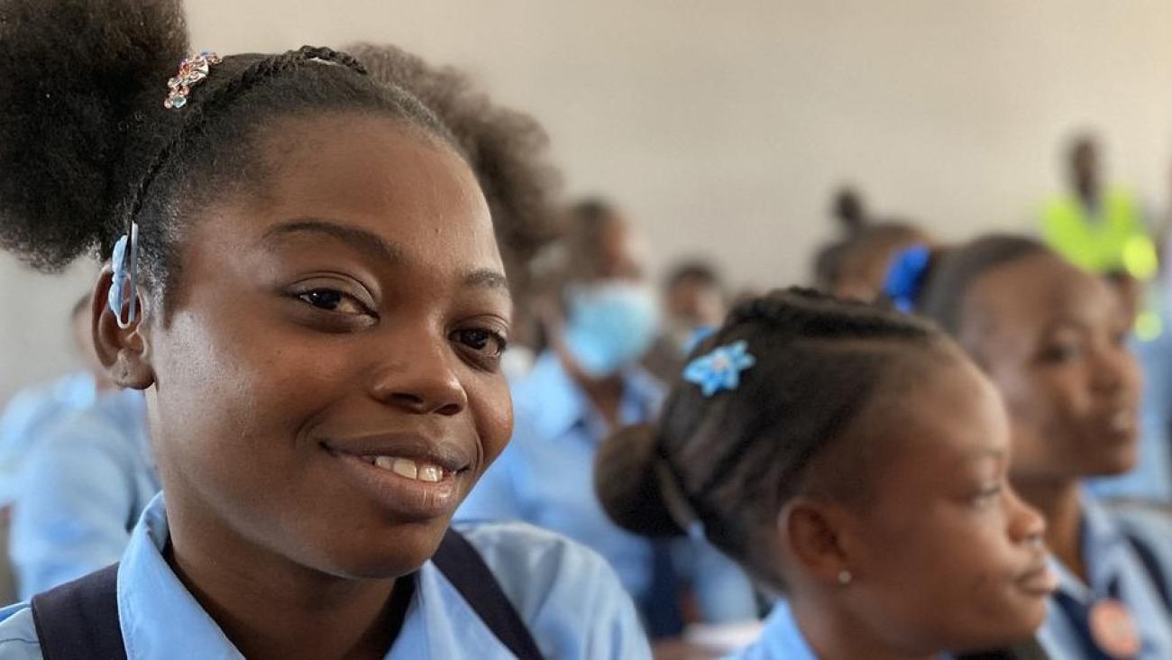Gros plan sur une jeune fille haïtienne en uniforme scolaire souriant à la caméra tandis que d'autres élèves, en arrière-plan, regardent en direction de l'enseignant.
