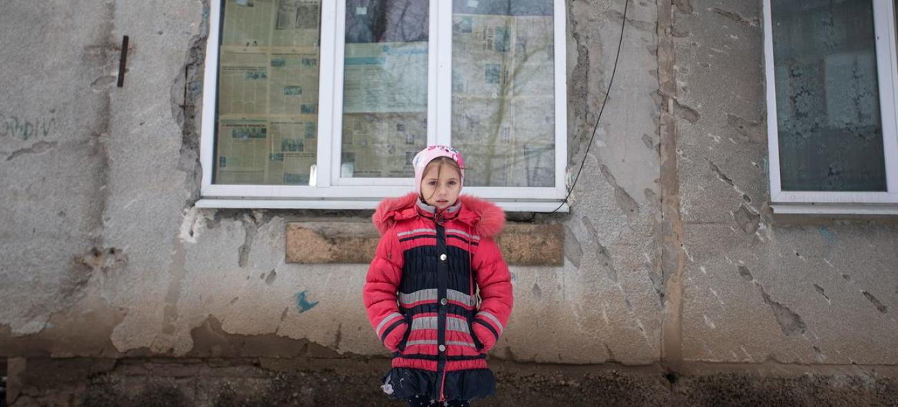 Una niña de nueve años, con un abrigo, se encuentra frente a la fachada de su casa, la cual ha sido dañada por el conflicto, en el este de Ucrania.