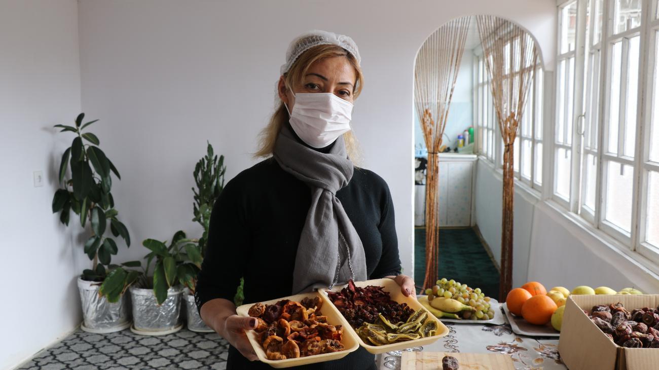 Une femme portant un masque de protection respiratoire blanc se tient debout près d’une table ou sont posés des fruits, un récipient de nourriture à emporter dans les mains.