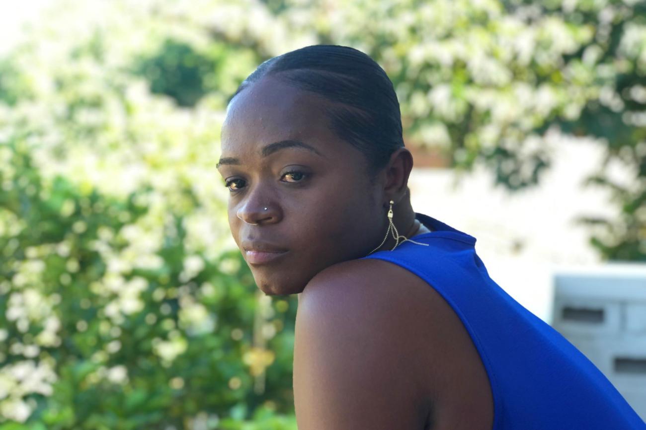 Une femme haïtienne en débardeur bleu penchée sur un balcon a le regard tourné vers sa gauche.
