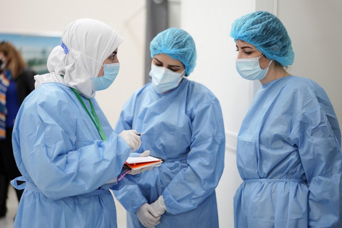 Trois professionnelles de santé portant chacune une blouse médicale, une charlotte et un masque de protection respiratoire discutent dans l’enceinte d’une établissement de santé.