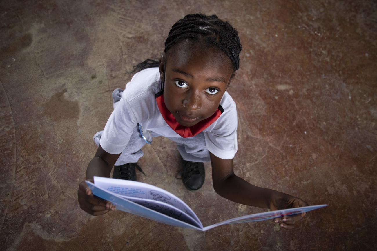 Une petite fille tient un livre ouvert entre les mains et lève la tête pour regarder l’objectif de la caméra.