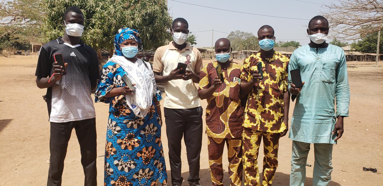 Un groupe de personnes portant un masque de protection respiratoire se tiennent debout, en extérieur, et montrent leur téléphone portable à la caméra.
