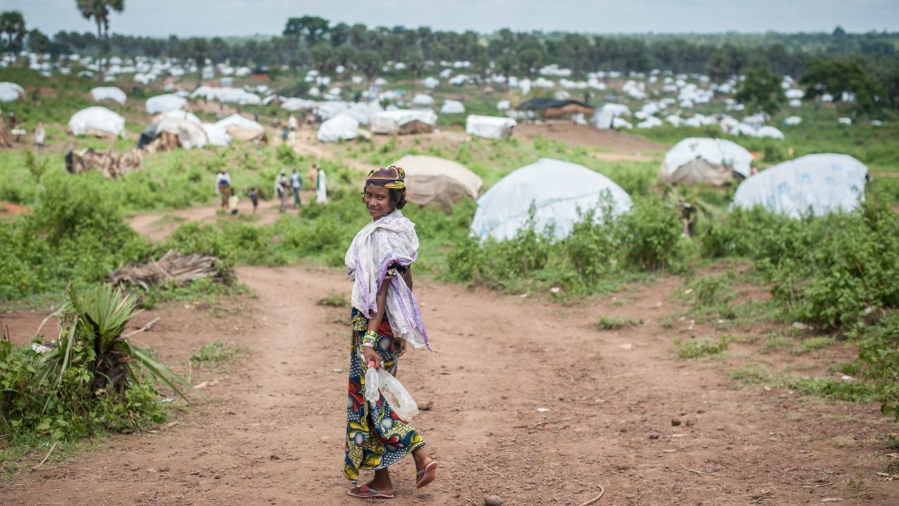 A woman turns back and smiles at the camera as she walks towards a green patch, with tents dotting the horizon.