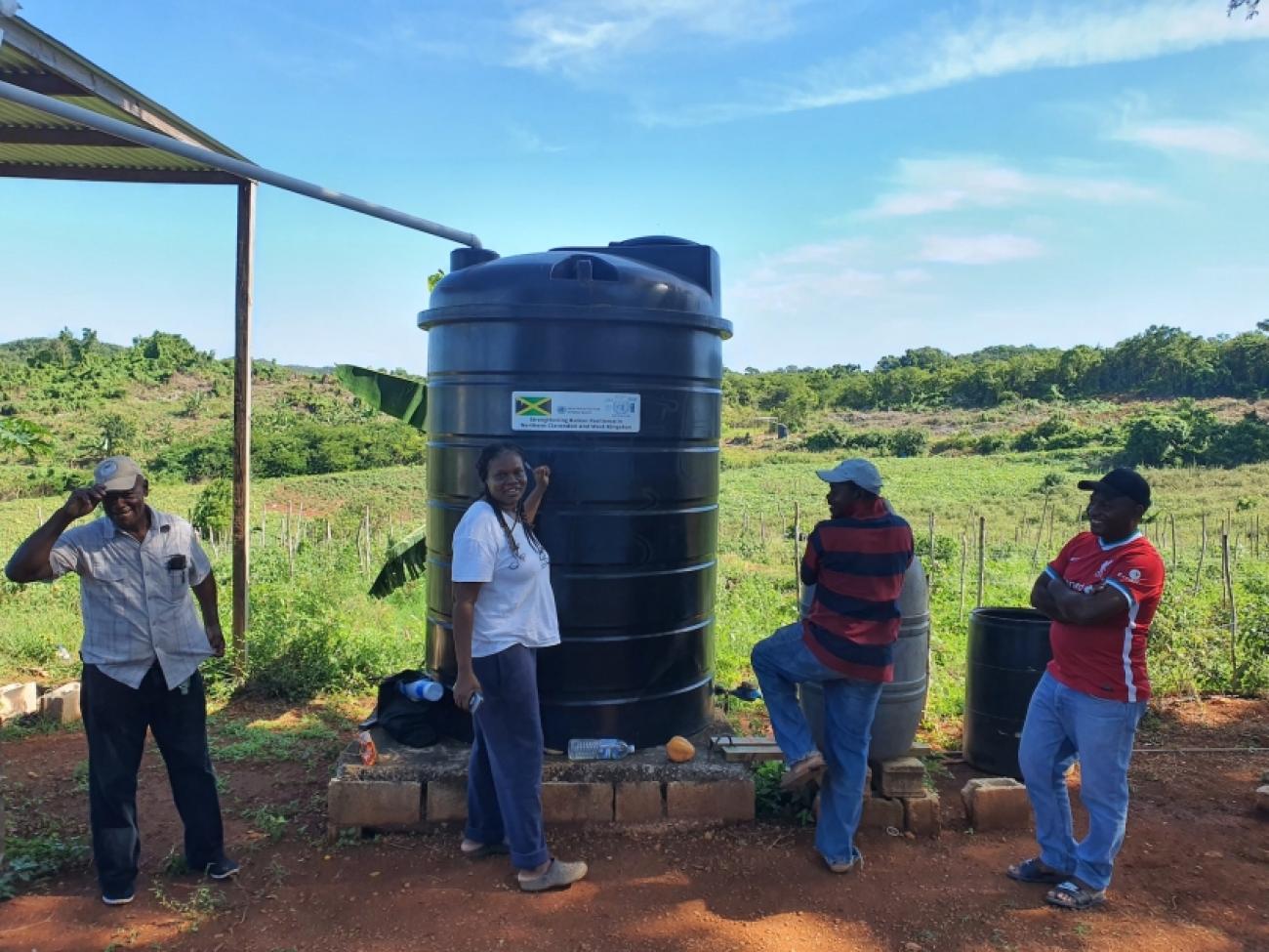 En Jamaïque, une femme et trois hommes se tiennent devant un réservoir d'eau installé dans un champ pour atténuer l'impact des sécheresses sur les hauts plateaux du pays.