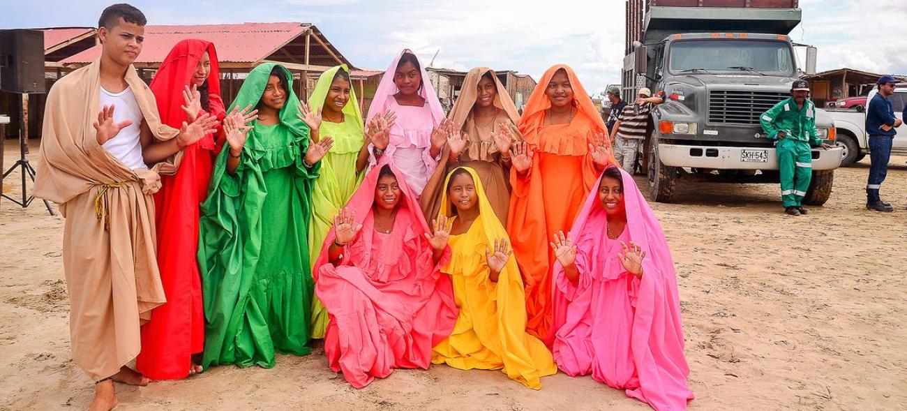 Youth in colorful dresses stand in a semi-circle against a tan-colored, sandy backdrop.