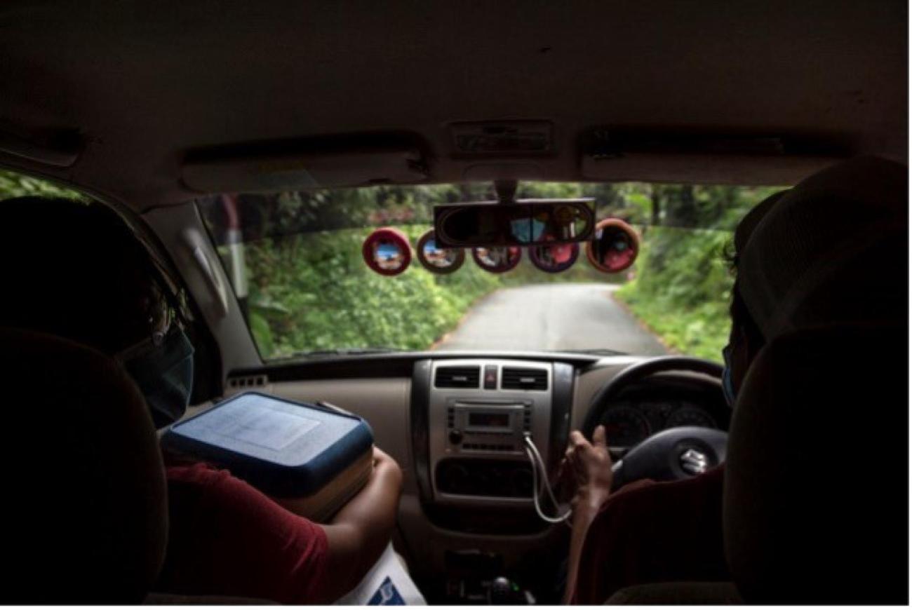 An illustrative photo from the backseat of a jeep in a green terrain, with one passenger carrying a box of what appears to be vaccines.
