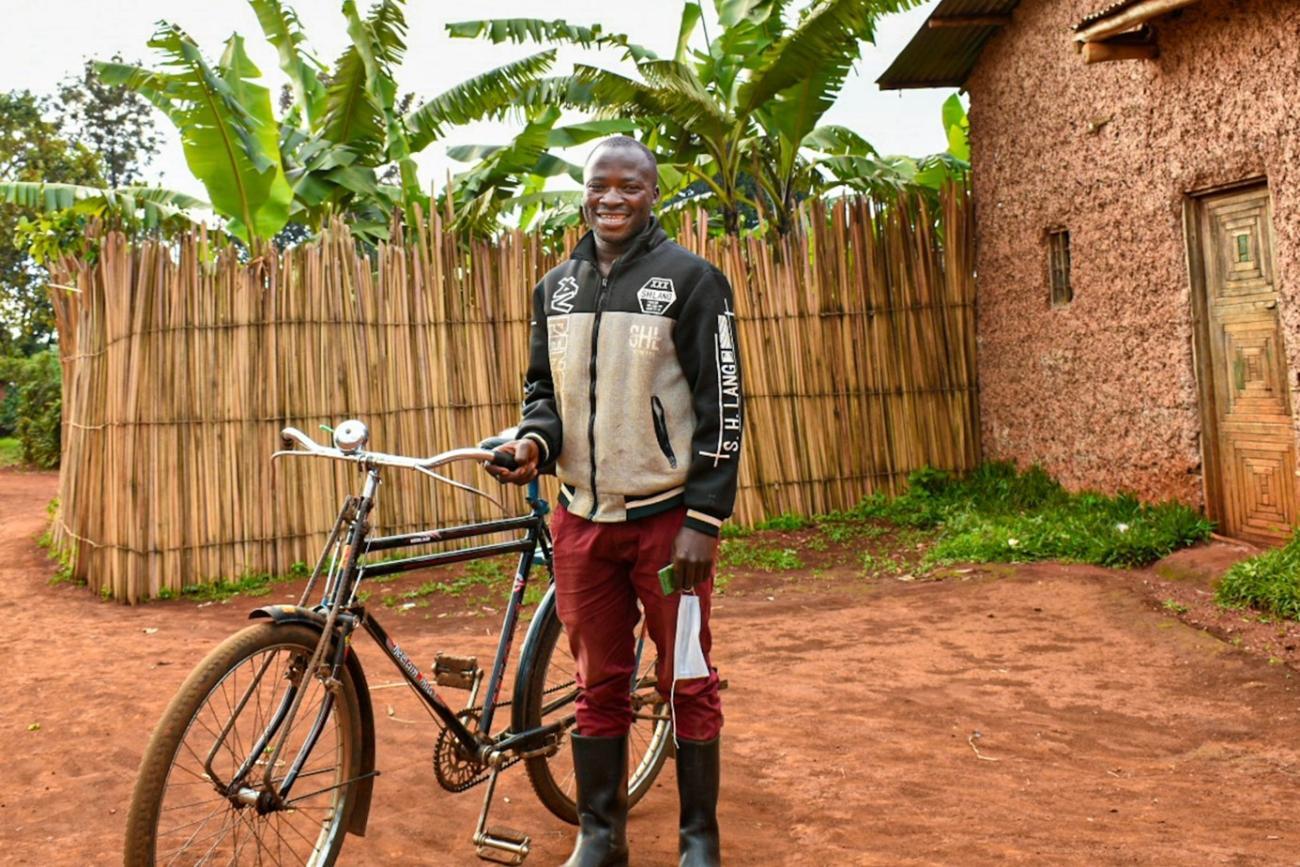 A man dressed in red pants stands by his bicycle outside a modest house lined with reeds and palm trees and smiles at the camera