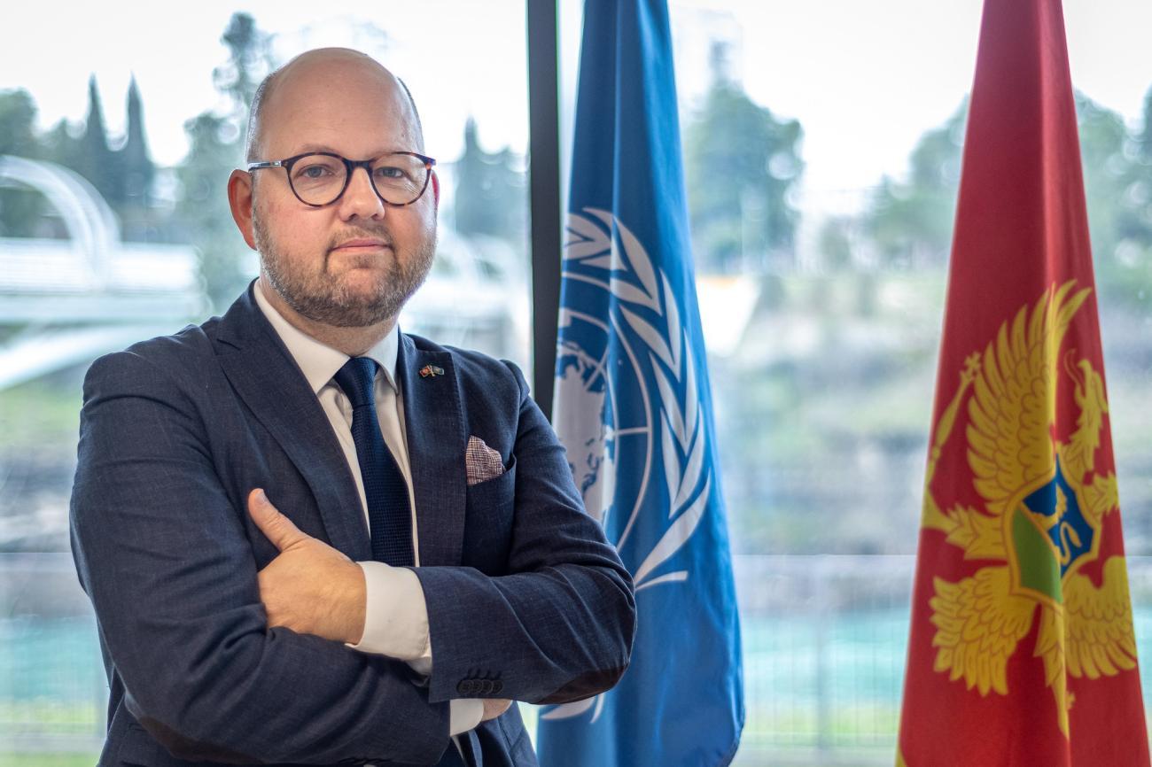 A man in a suit standing next to the flags of United Nations and Montenegro.