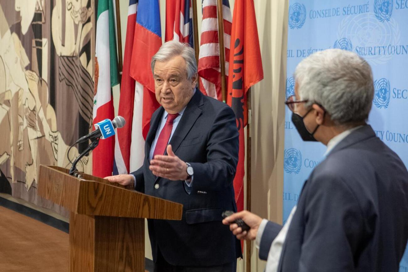 Un hombre habla por un micrófono con banderas detrás de él. Es el Secretario General de la ONU, durante una sesión informativa para los medios de comunicación.
