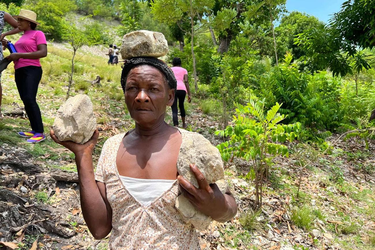 A Haitian woman stands facing the camera, in the middle of a steep terrain littered with shrubs. She holds a large stone in each hand.