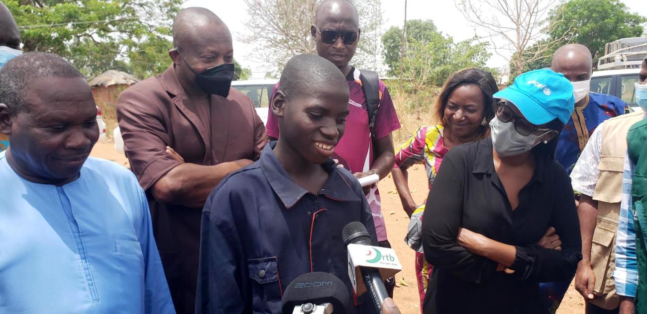 Un jeune adolescent béninois vêtu d'une blouse de mécanicien et entouré de plusieurs hommes et femmes répond aux micro de plusieurs journalistes en souriant. 