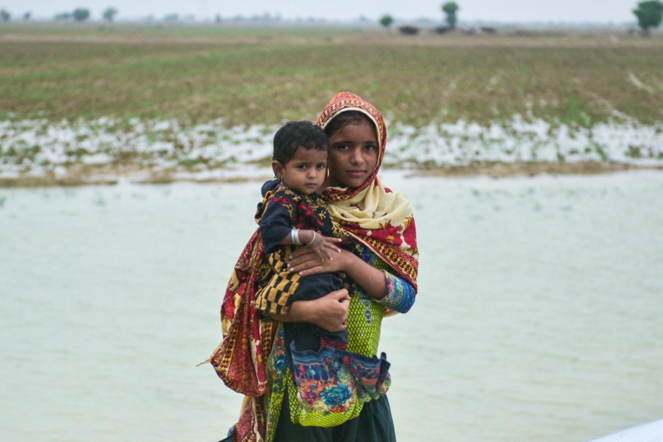 A girl in scarf holds a toddler in her arms in front of a pool of water collected on a field, against a grey sky. 