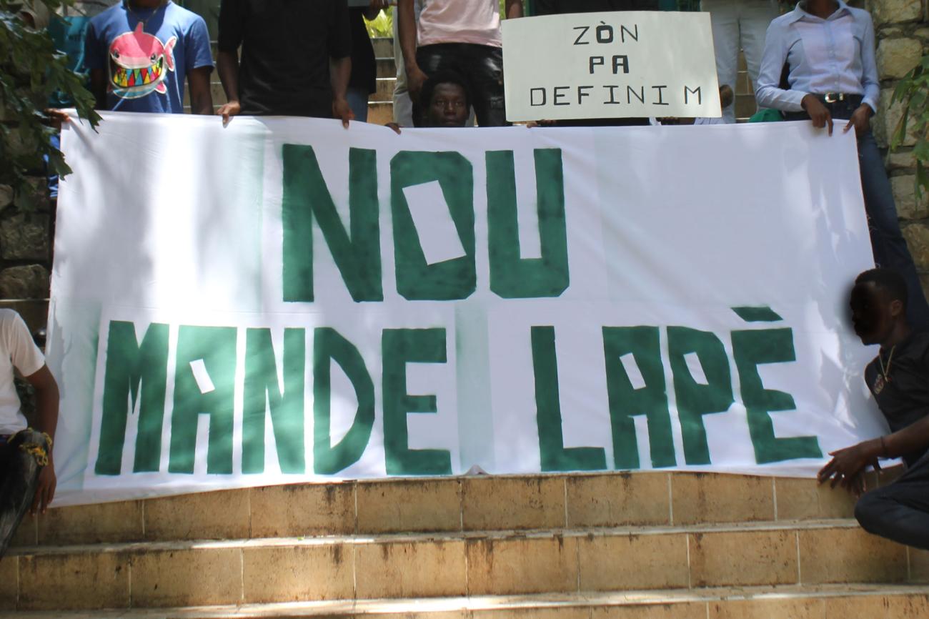 Un plano detalle de una gran pancarta con grandes letras verdes, que es sostenida por activistas juveniles quienes exigen paz a través de un eslogan escrito en criollo.