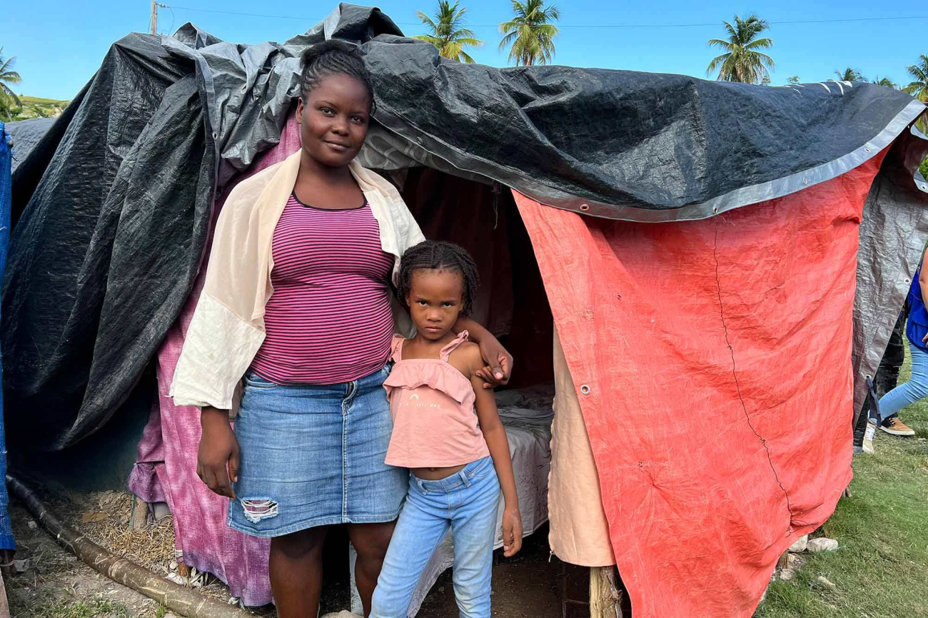 A young woman and her daughter stand in front of a temporary shelter covered with canvas and stare into the camera's lens on a sunny day in Haiti.
