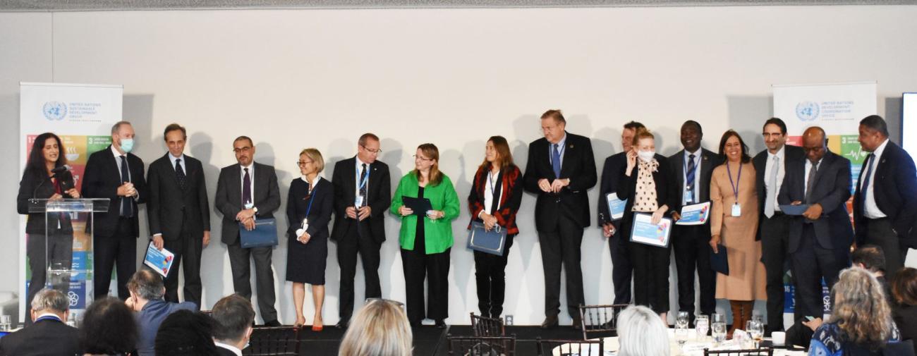 Un grupo de dieciséis personas en traje de negocios, de pie uno al lado del otro, aplaudiendo y felicitándose, en un escenario durante la ceremonia de entrega de los Premios a los Informes Anuales de Resultados de los equipos de las Naciones Unidas.