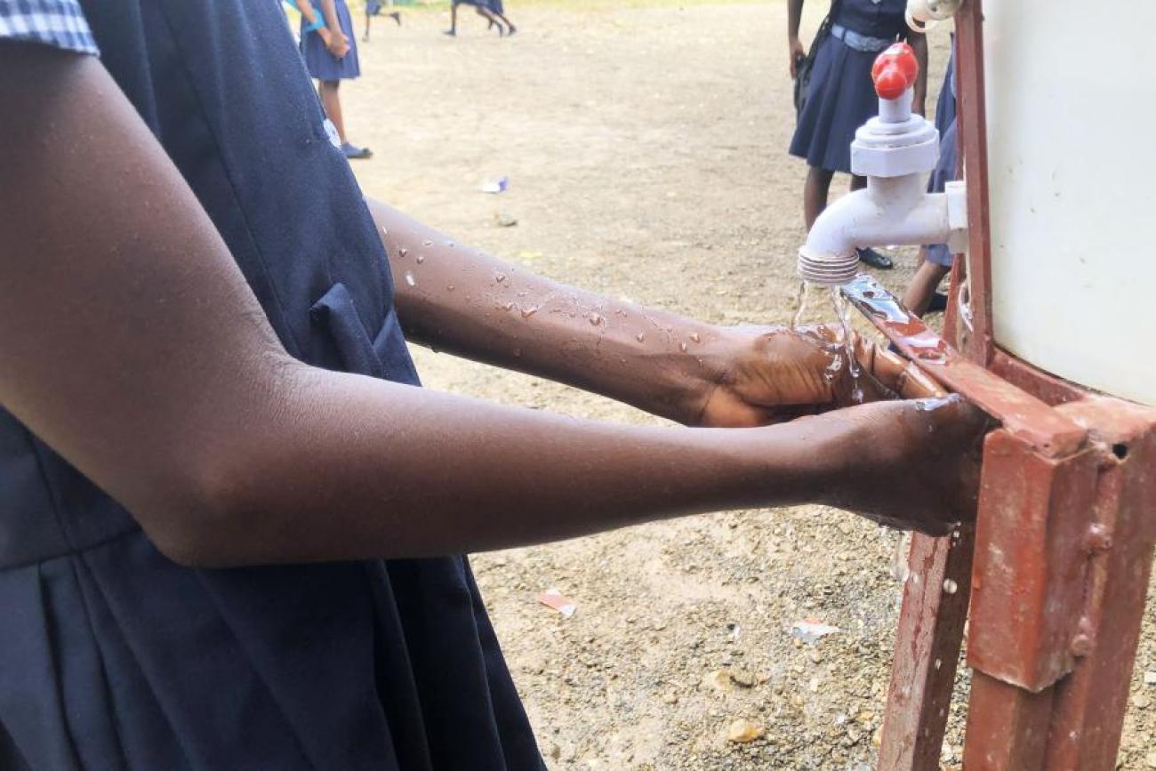 Una niña con lo que parece ser un uniforme escolar azul oscuro se lava las manos con jabón en un espacio exterior con suelo de tierra.
