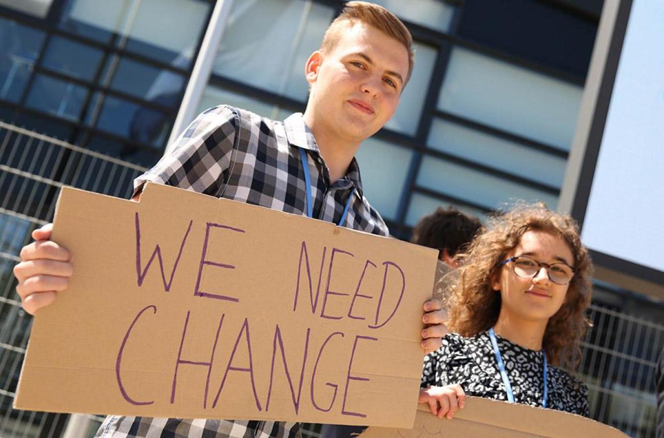 Un hombre joven sostiene una cartulina con el texto "WE NEED CHANGE" ("NECESITAMOS CAMBIO", en inglés) escrito en ella, y a su lado, una chica sostiene otra pancarta, cuyo texto es ilegible.
