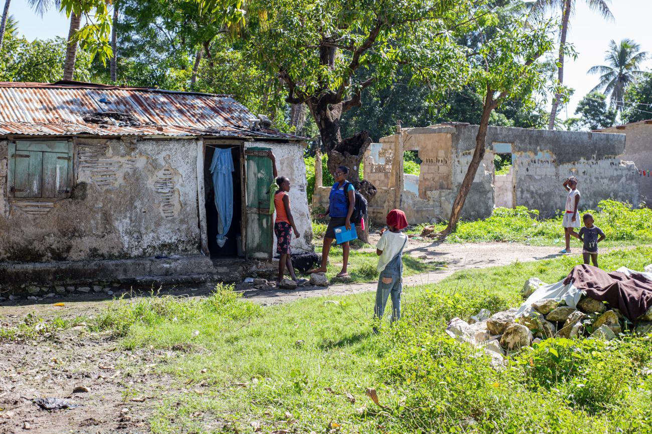 Dos mujeres jóvenes conversan a la entrada de una vieja casa en ruinas rodeada de árboles. Varios niños y niñas juegan cerca.