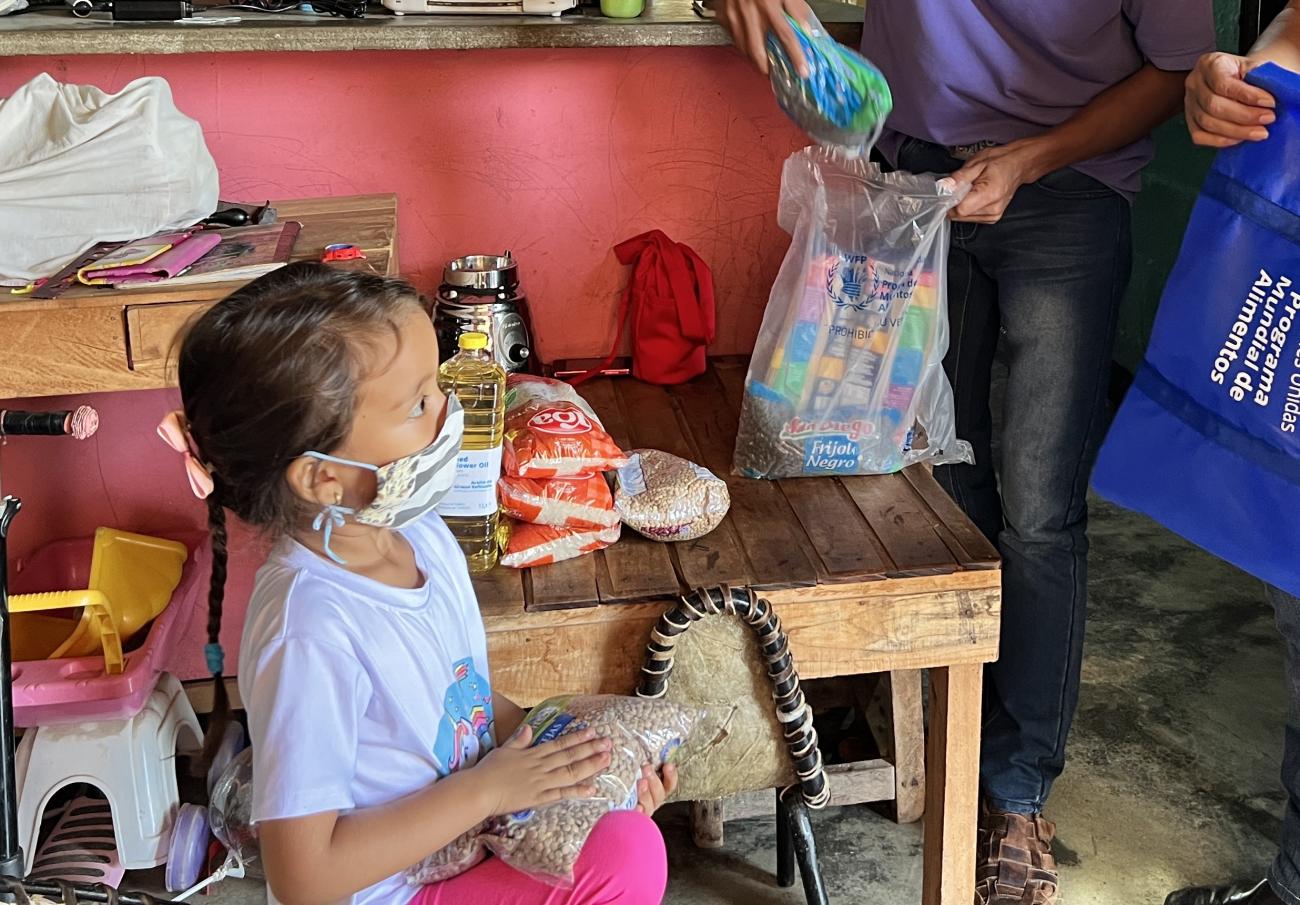 A small girl with a facemask holds a bag of beans while looking at two adults, and in the background there is a table with food on it.