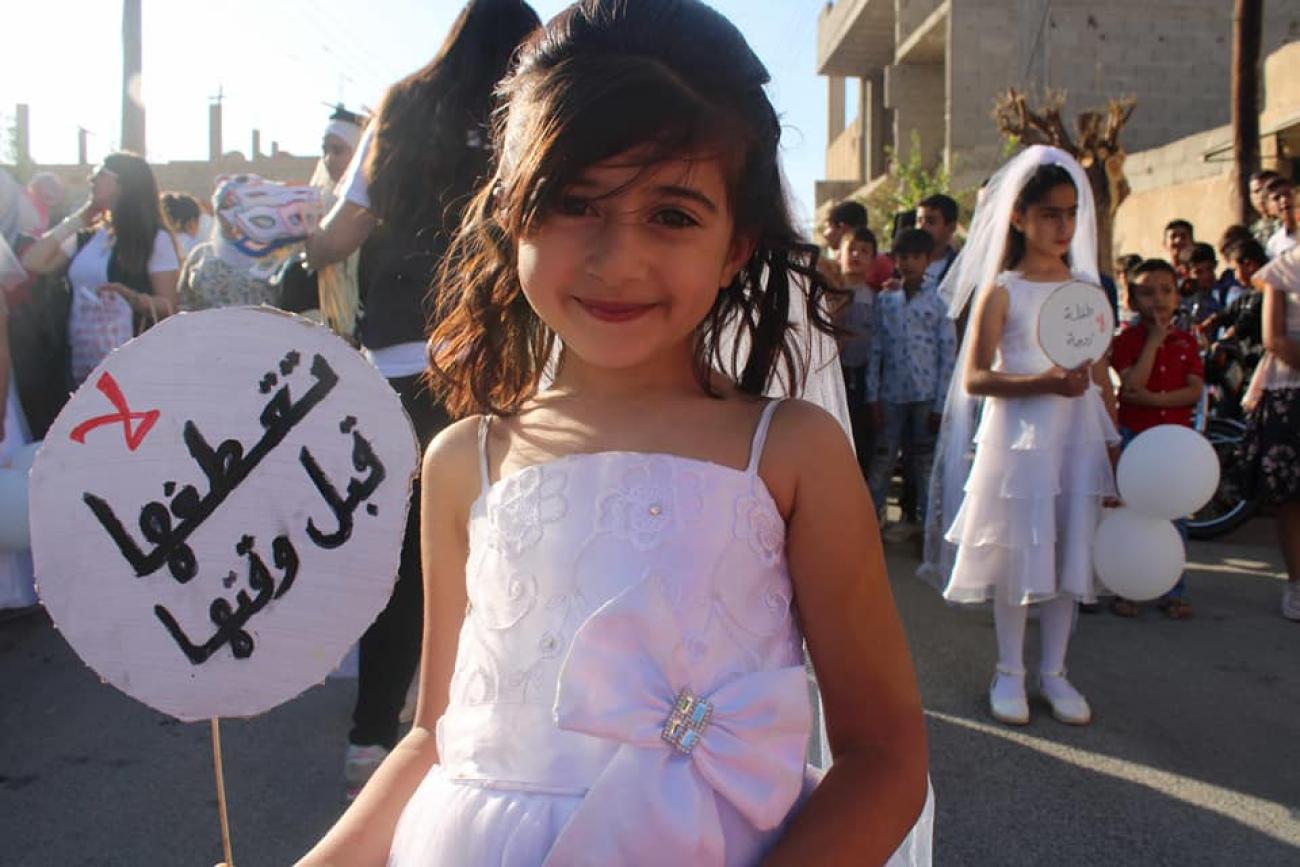 Young girl wearing a wedding dress and carrying a sign that says: don't marry her before her time.