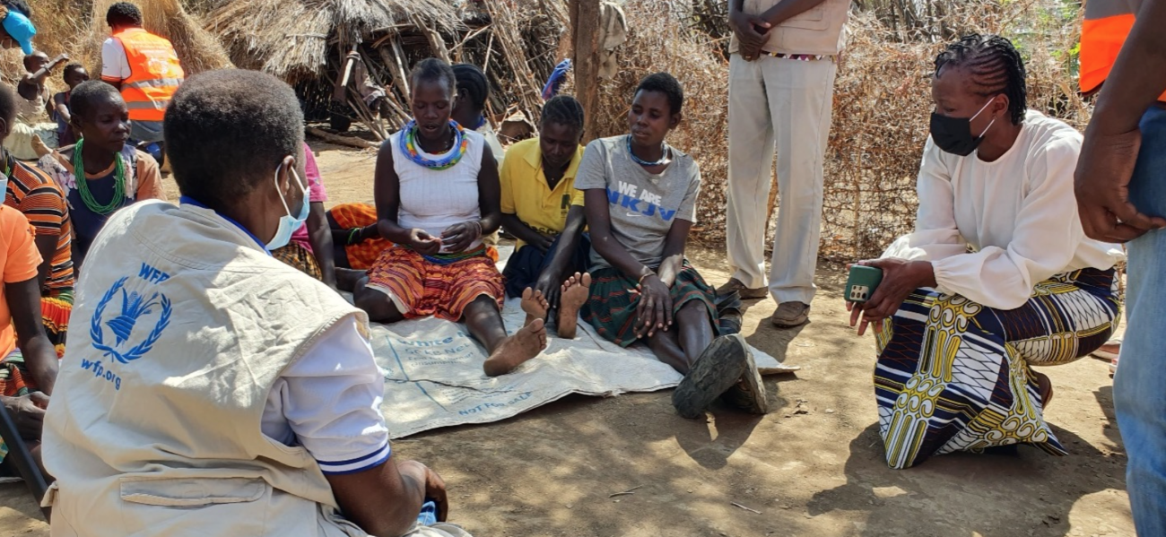 A group of people sit in a circle on the tan ground and discuss nutrition in Uganda