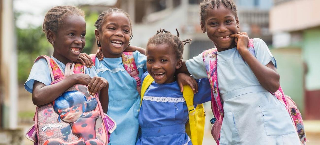 School-age girls in braids and colorful t-shirts and backpacks giggle and embrace, and face the camera.