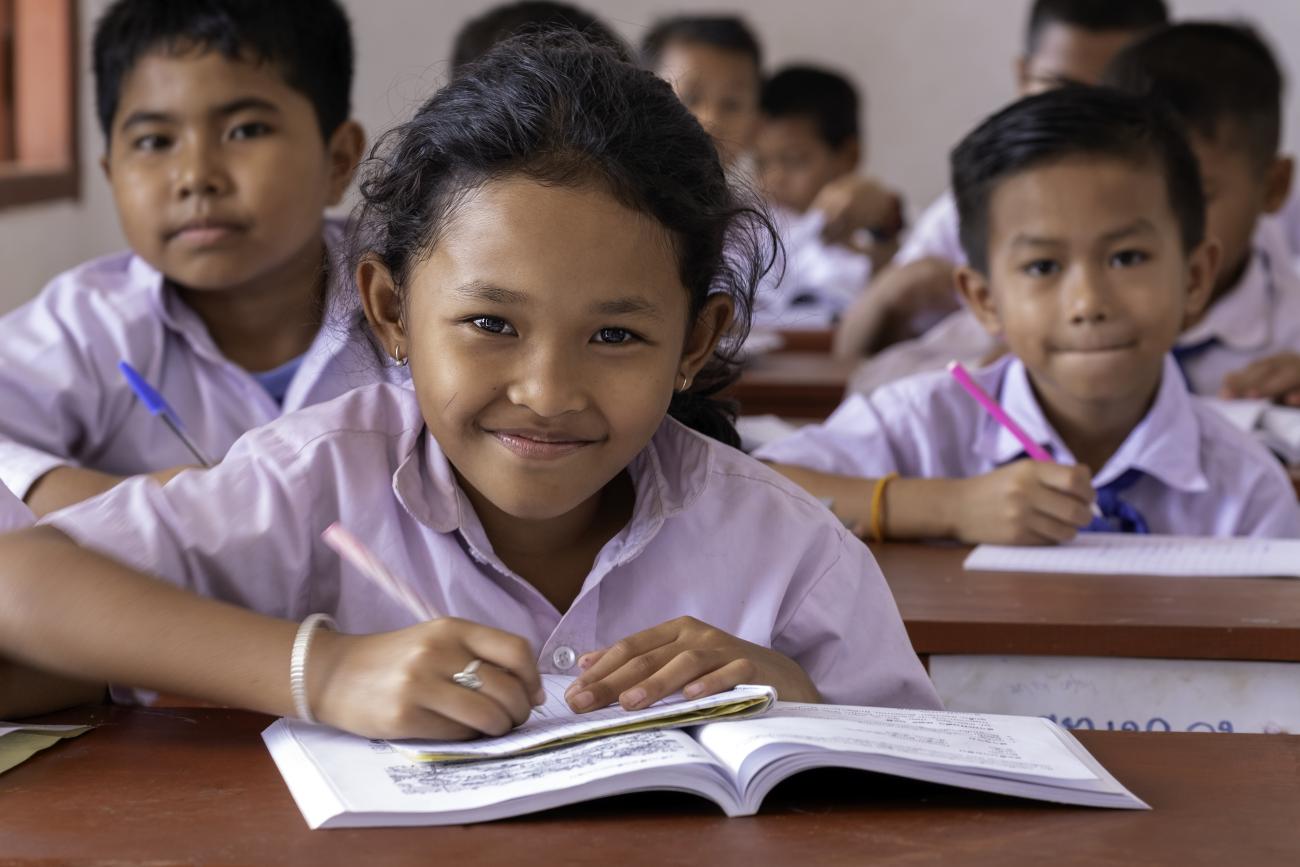 Children in white school uniforms sitting at a desk with books open smiling to camera