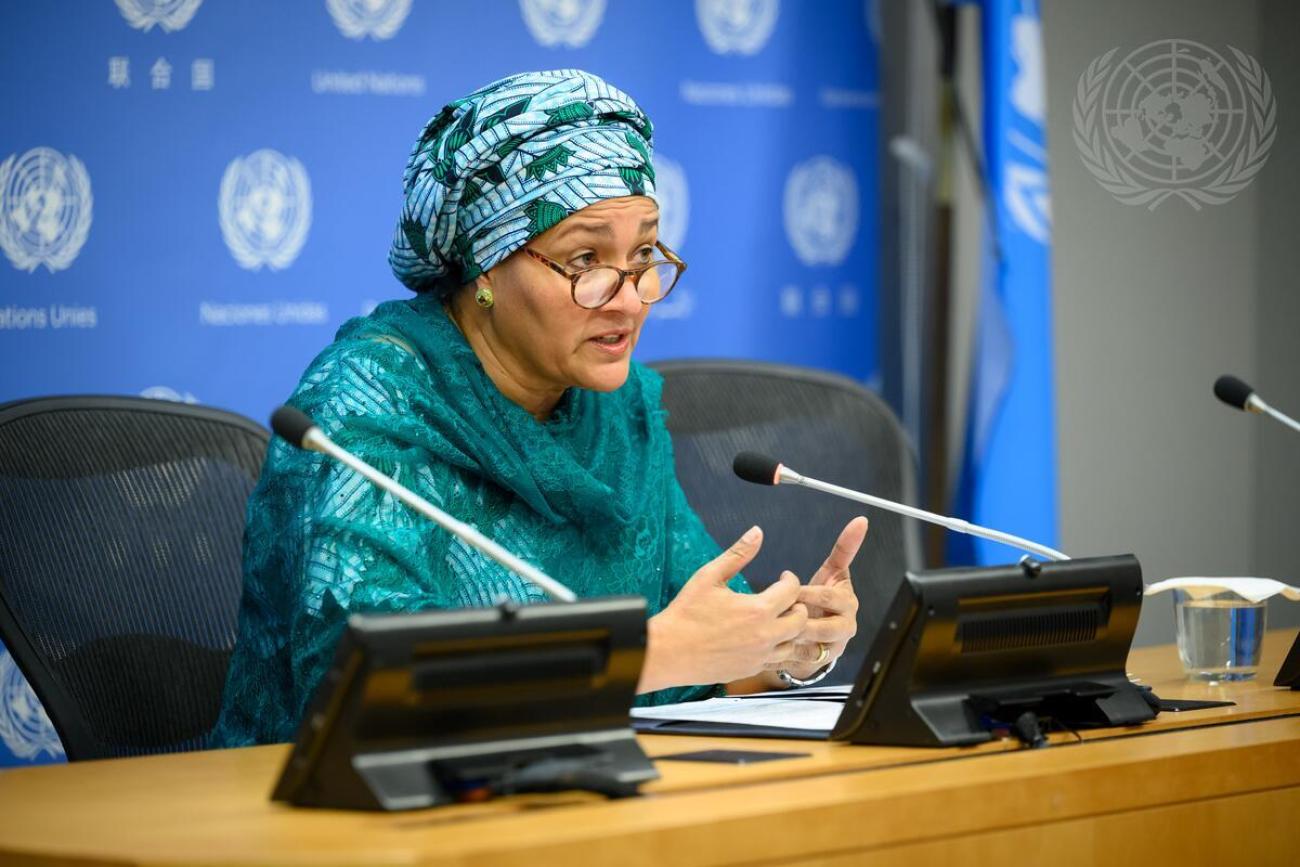 Woman in a green dress and headscarf speaking at a podium into a microphone against a background with the UN logo