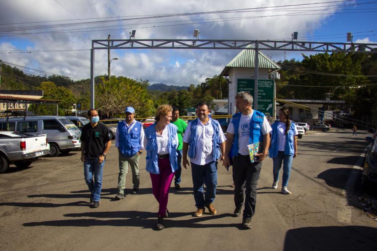 A group of people in different white and blue shirts and vests walk and talk through a grey road where cars are parked