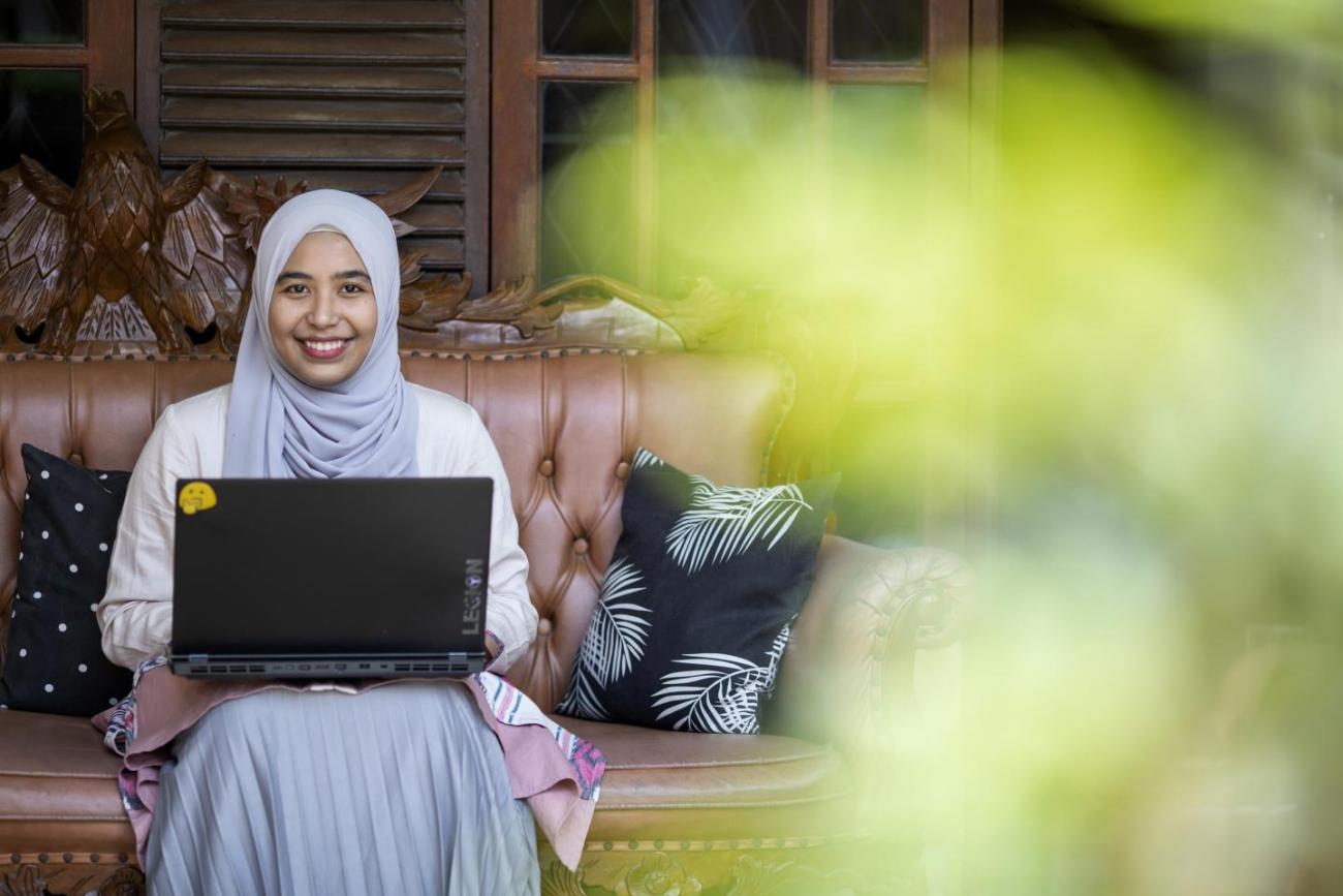 Woman in a white and grey headscarf sits on a bench with a laptop in her lap