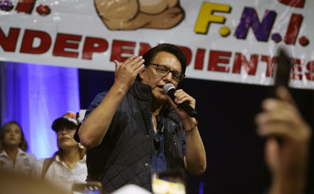 A man at a rally speaks into a microphone with a banner behind him.