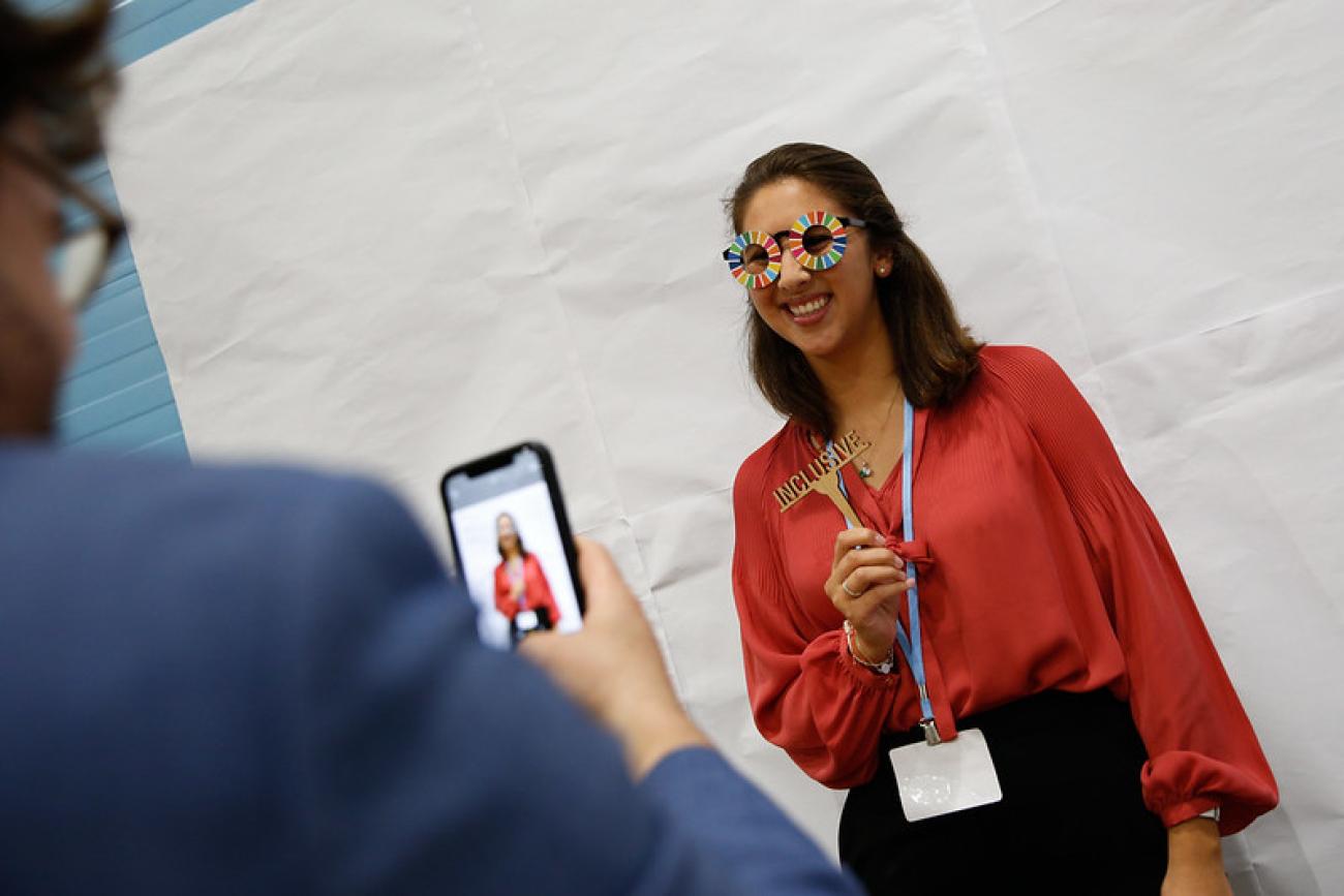 A woman in a brown shirt with glasses shaped like the UN SDGs smiles as someone else takes a photo of her on their mobile phone