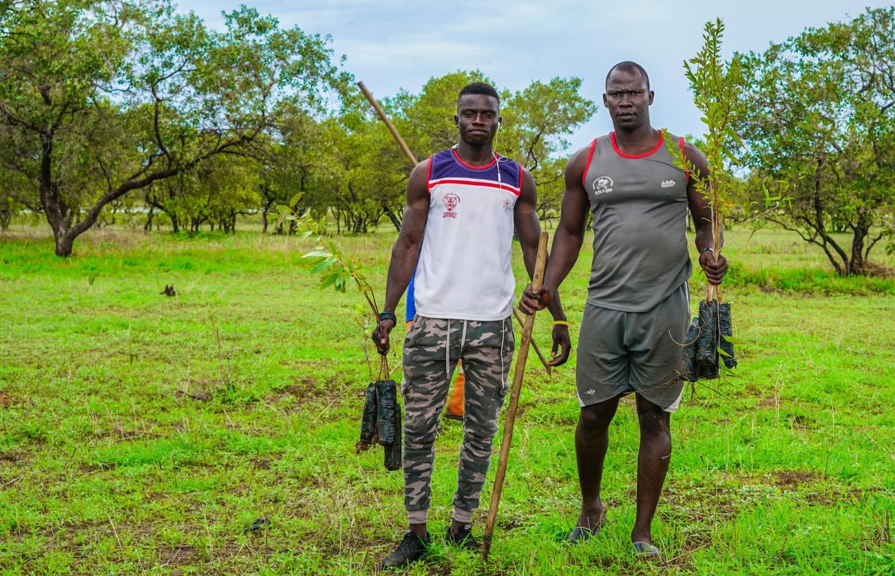 Two men, one in a white shirt and the second in a grey shirt, stand in a green field holding planting tools 