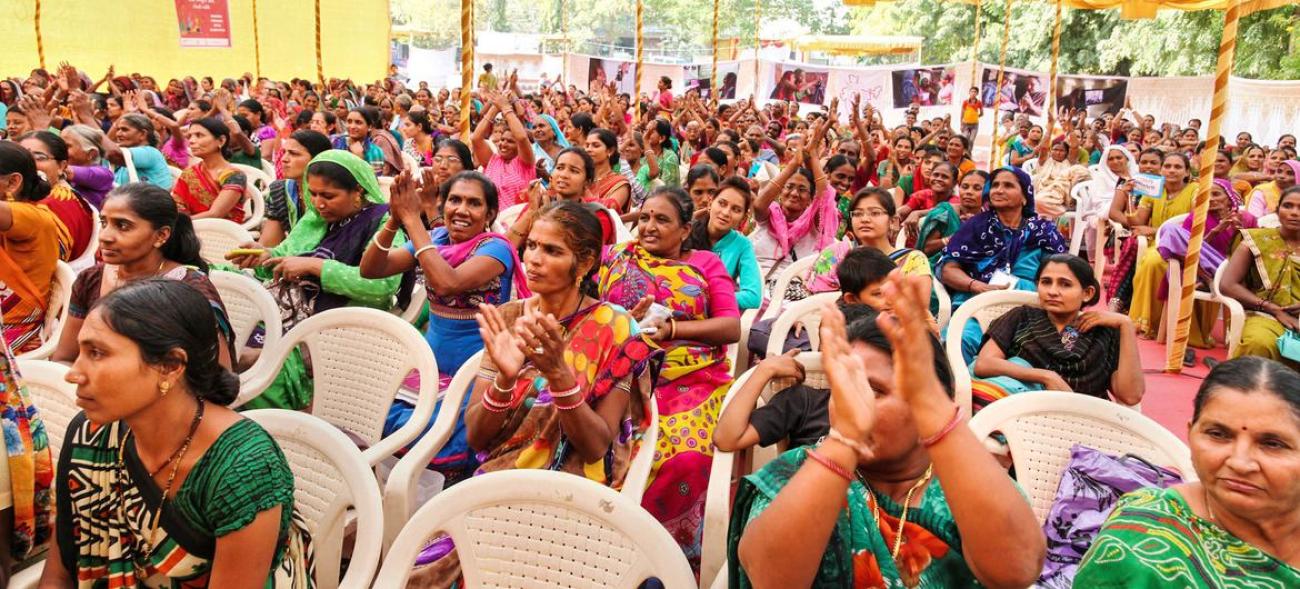 مجموعة من النساء يرتدين ملابس جنوب آسيا التقليدية الملونة يجلسن على الكراسي في اجتماع حاشد أو حدث.
