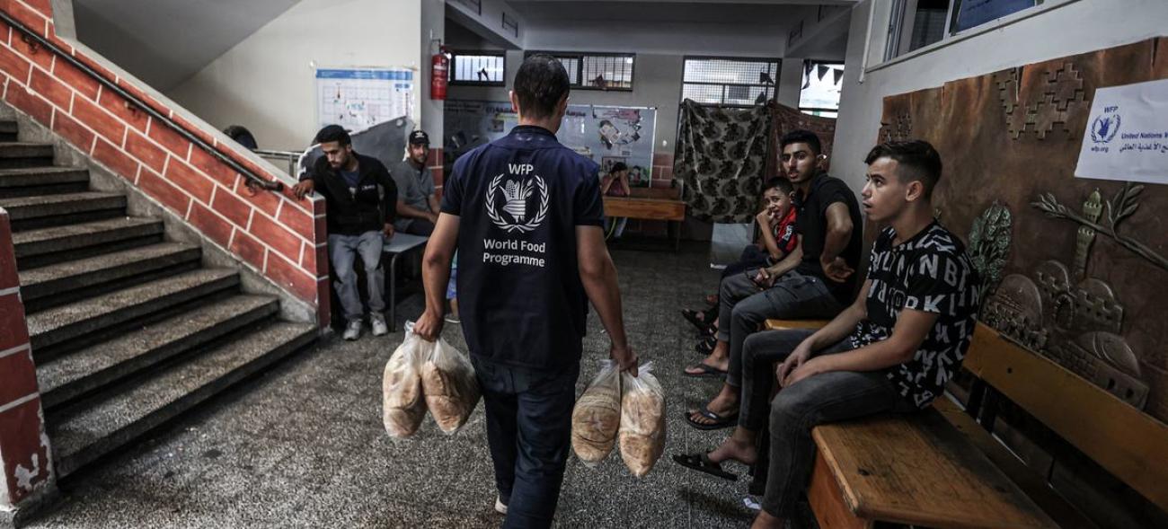 A WFP humanitarian worker in a black shirt carries bags of grain into a UNWRA school in Gaza where communities are seeking refuge.