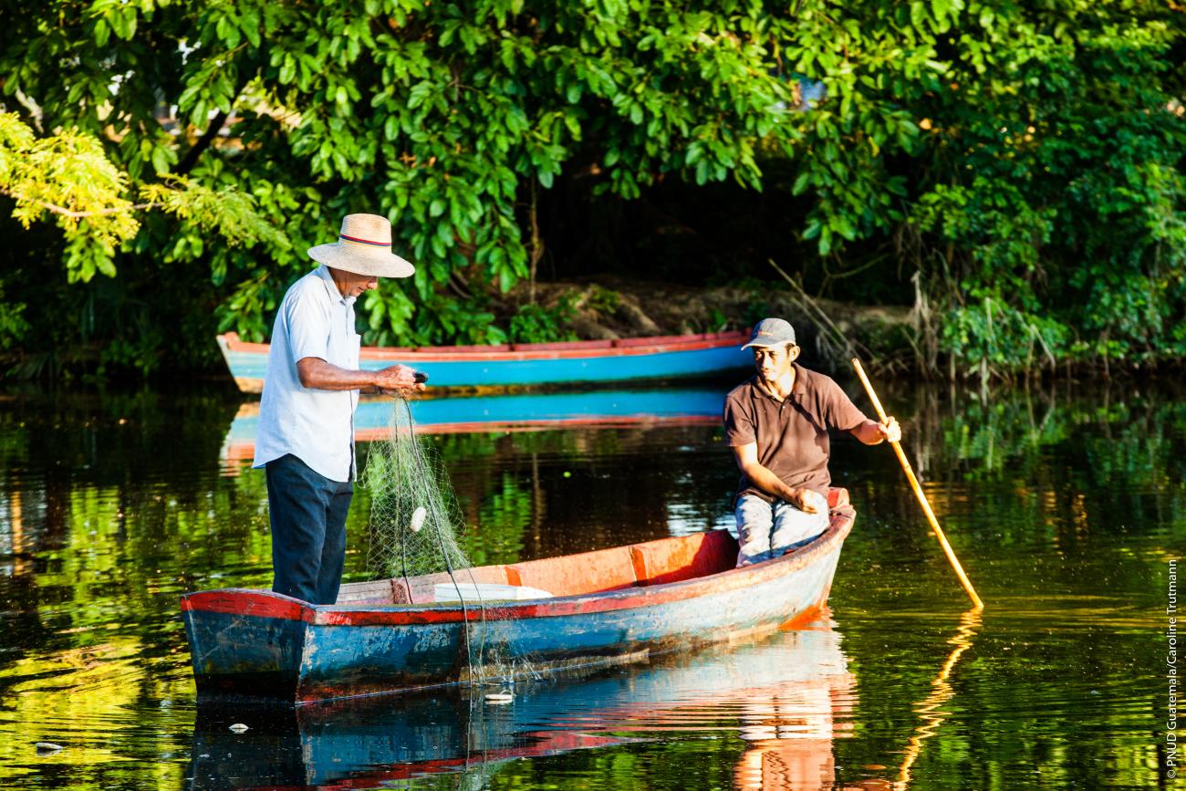 Two fisherman on a small boat or canoe, on a body of water surrounded by greenery. 