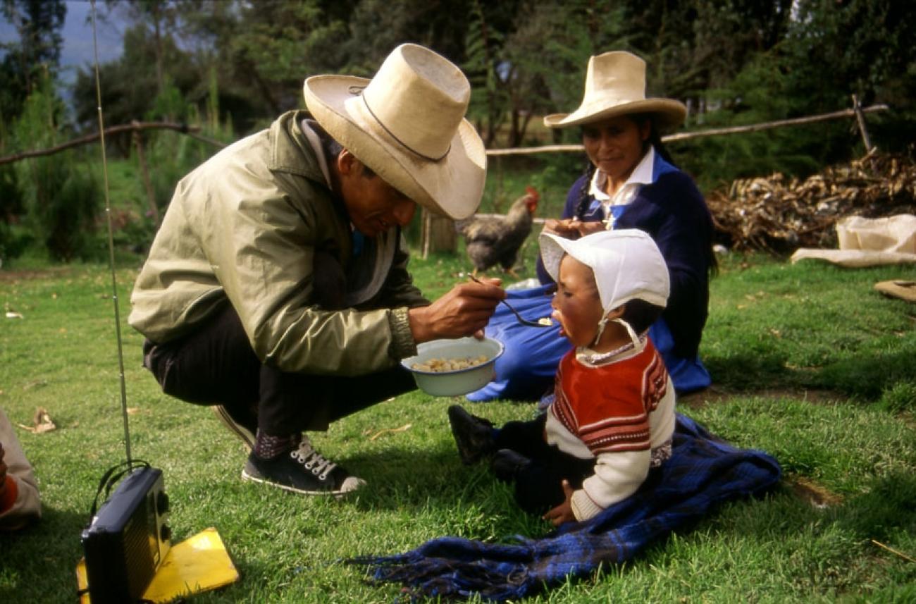 A man in a hat feeds an infant while a woman sits by the infant on a grass area in the sun.
