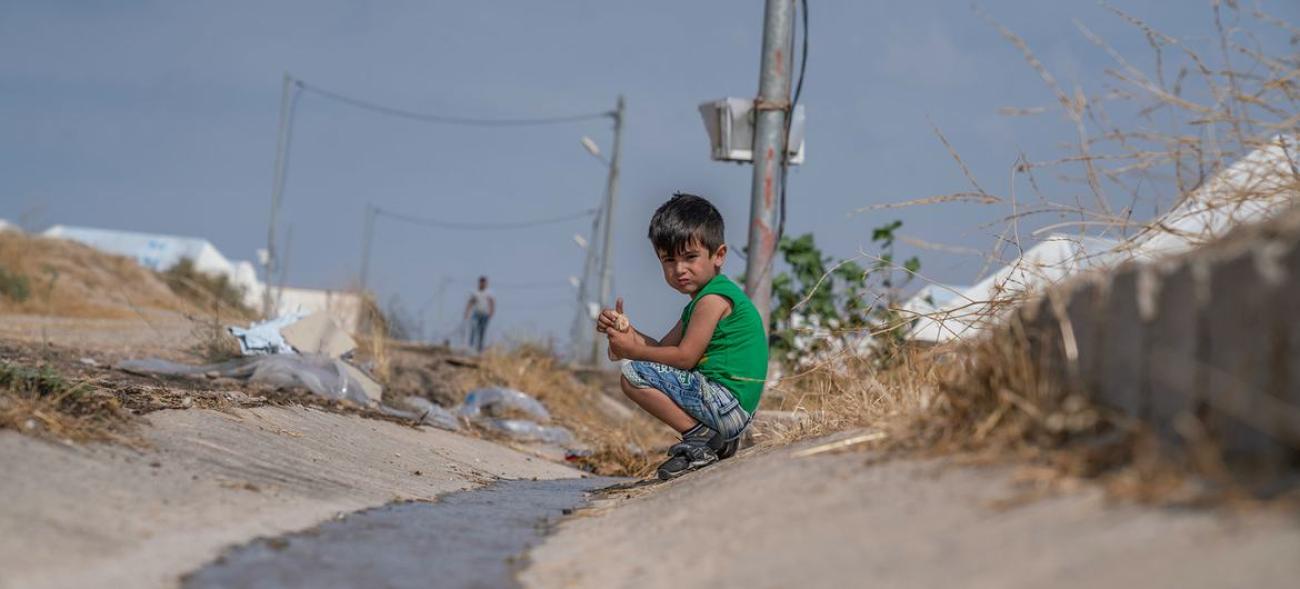 A little boy in a green shirt and blue pants, a Syrian refugee, kneels on the sound of a road in Iraq.