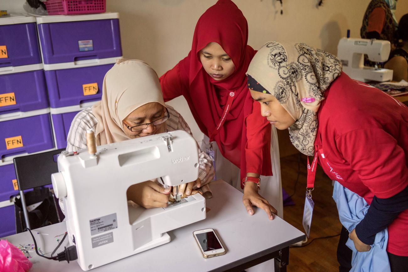 three women sit around a sewing machine wearing headscarves.